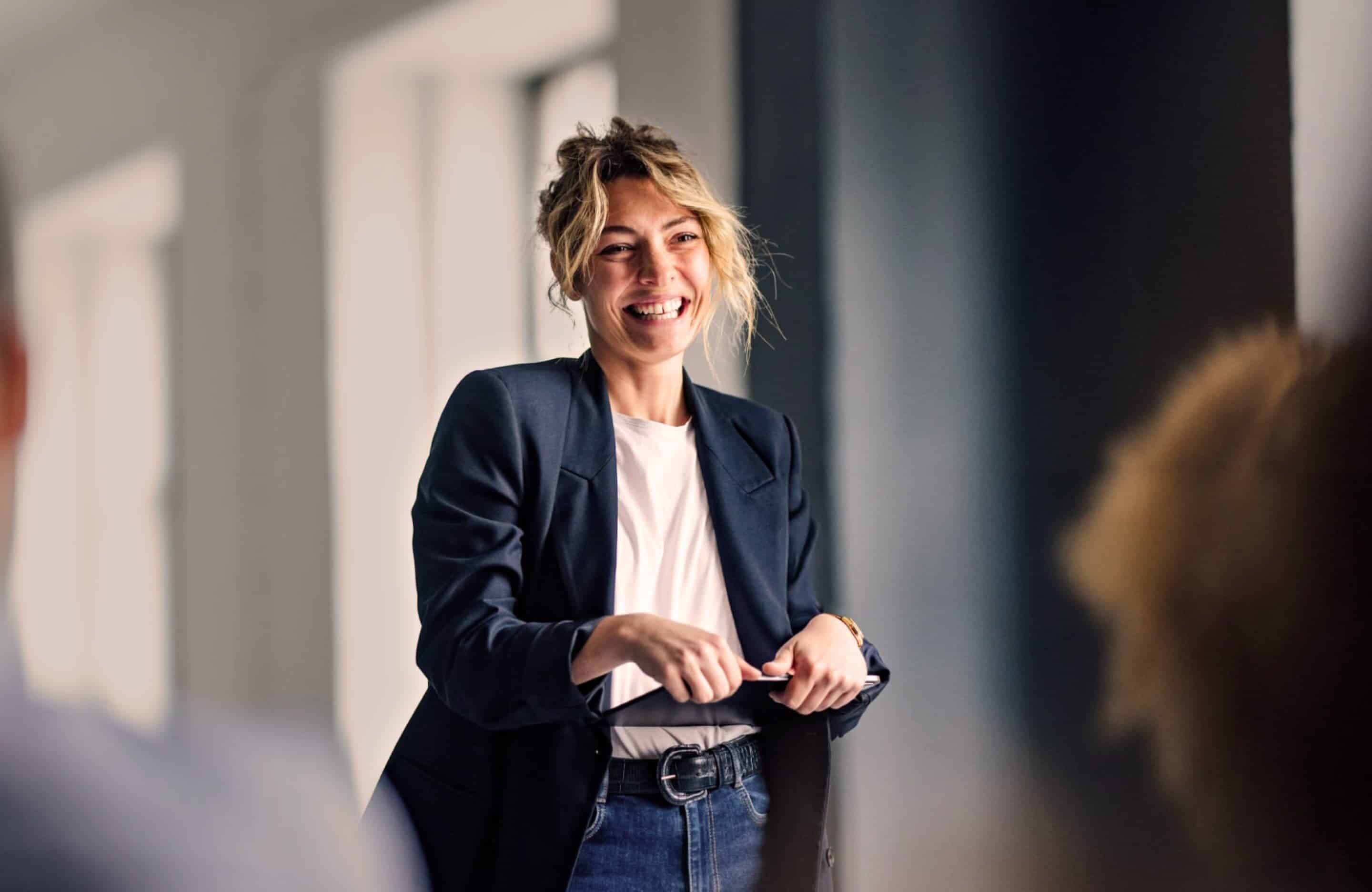 Professional woman in navy blue jacket and jeans holds iPad and smiles at workplace